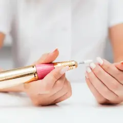 Dłonie z białymi paznokciami trzymają różowo-złotą frezarkę. Dowiedz się, jak używać frezarki do paznokci, aby uzyskać perfekcyjny manicure.