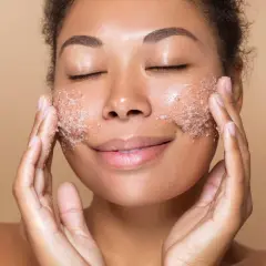 Kobieta z zamkniętymi oczami delikatnie wmasowuje peeling do twarzy, ciesząc się chwilą relaksu i odnowy skóry.