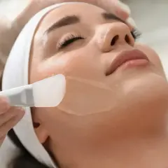 Kosmetyczka aplikuje żelowy peeling na twarz klientki. To pierwszy krok do gładkiej skóry.