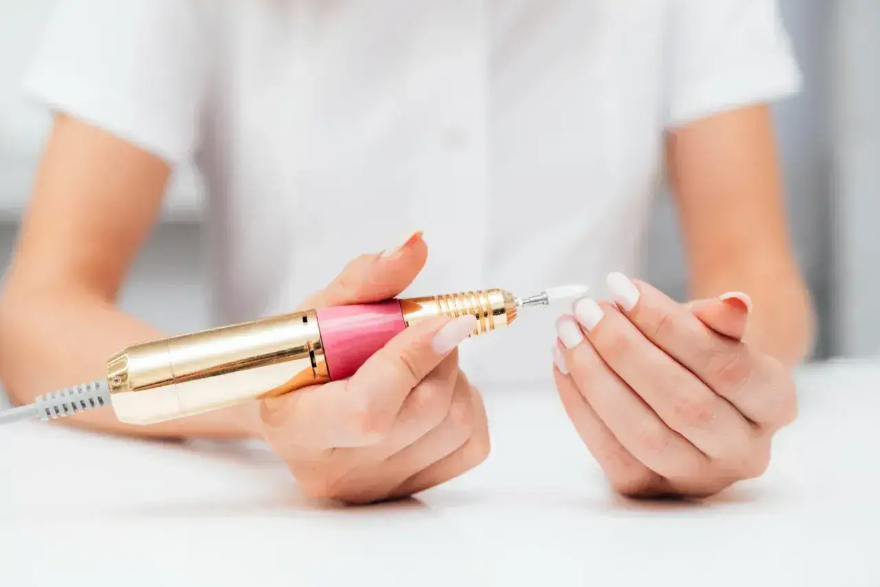 Dłonie z białymi paznokciami trzymają różowo-złotą frezarkę. Dowiedz się, jak używać frezarki do paznokci, aby uzyskać perfekcyjny manicure.