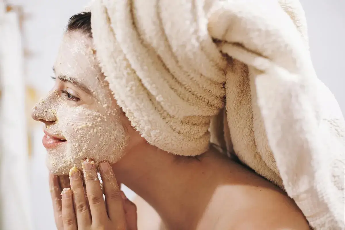 Kobieta z ręcznikiem na głowie nakłada na twarz domowy peeling. Delikatne drobinki złuszczają naskórek, przygotowując skórę na dalsze zabiegi.