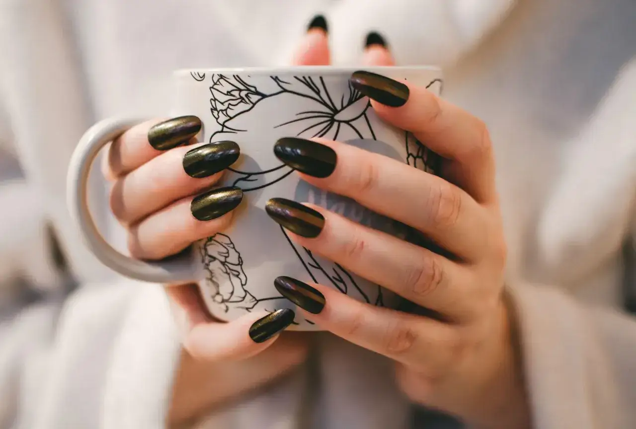 Dłonie z ciemnym, błyszczącym manicurem japońskim trzymają kubek z rysunkiem.